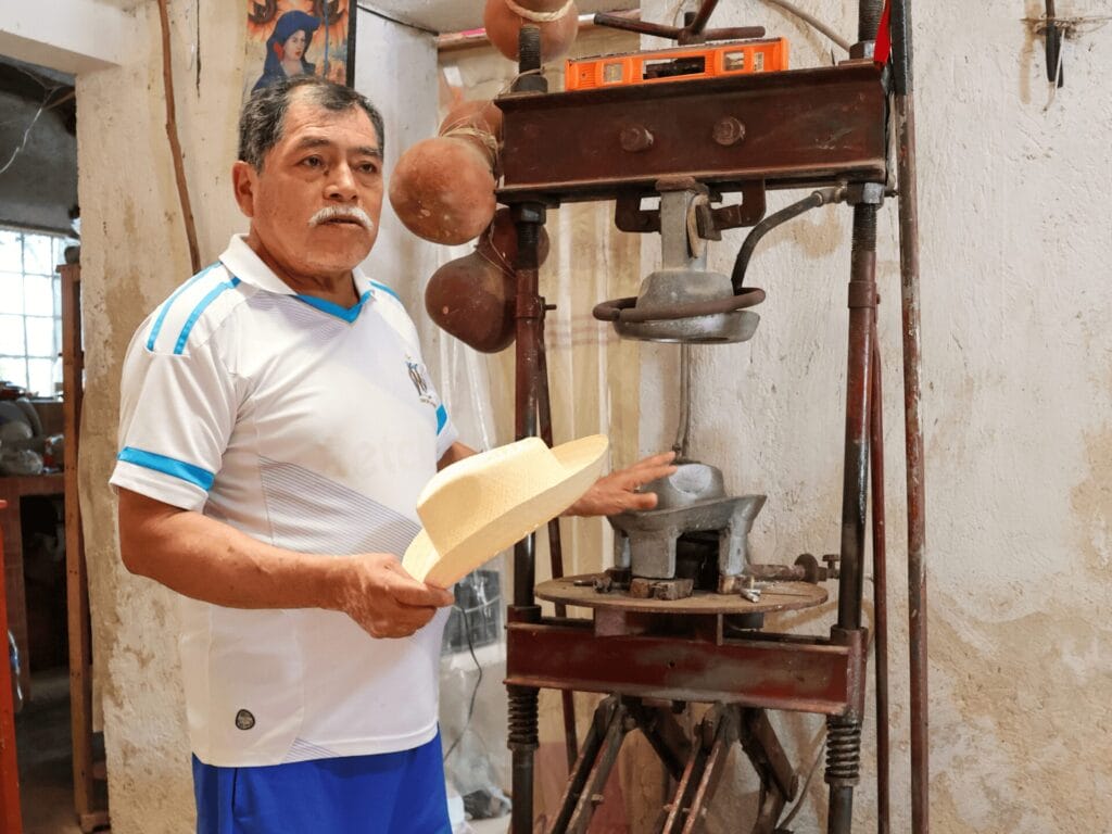 Ein mexikanischer Mann mit schwarzen Haaren und einem grauen Schnautz, der ein weisses T'Shirt und blaue Shorts trägt, steht neben einer Hutpresse. In der rechten Hand hält er einen naturfarbenen Strohhut, mit der linken Hand zeigt er auf die Hutpresse.
