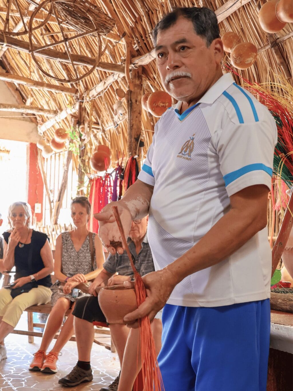 Ein mexikanischer Mann mit schwarzen Haaren und einem grauen Schnautz, der ein weisses T'Shirt und blaue Shorts trägt, hält einen Tontopf und gefärbte Strohhalme in der einen Hand und Färbepulver in der anderen Hand. Im Hintergrund sind zwei zuschauende Frauen zu sehen. Die Personen befinden sich in einer strohbedeckten Halle.